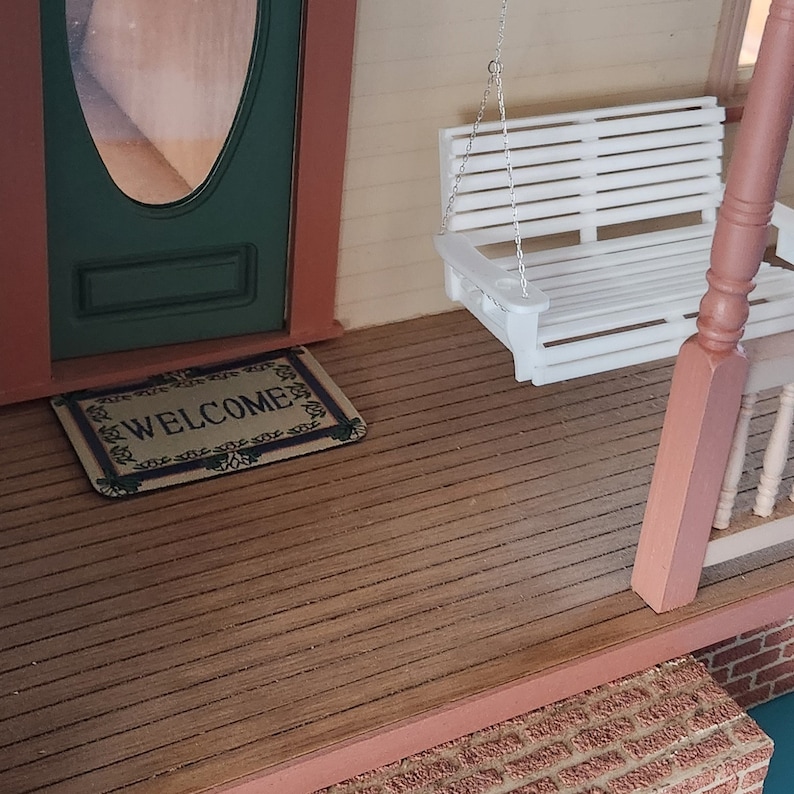 May include: A miniature dollhouse porch with a white swing hanging from the ceiling, a welcome mat on the floor, and a wooden floor with red trim.