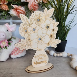 May include: A wooden floral arrangement with the text "Flowers for Mommy" engraved on the vase. The flowers are laser-cut with intricate details and small mirrored accents. The arrangement sits on an oval base.