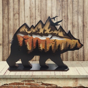 May include: A wooden bear silhouette with a layered mountain scene inside. The mountain scene is painted with shades of brown, orange, and yellow. The bear is black and has a flat base.