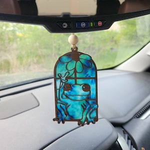 May include: A blue and green frog with a flower in its hand is inside a wooden frame. The frame is hanging from a rearview mirror in a car.