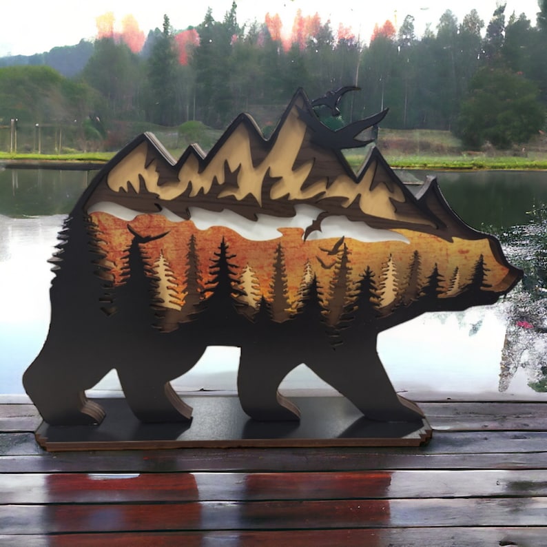 May include: A wooden bear silhouette with a layered design featuring a mountain range, a forest, and a sunset. The bear is standing on a wooden base.