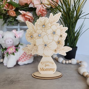 May include: A wooden floral bouquet decoration with the words "Flowers for Mommy" engraved on the vase. The bouquet features etched flower and leaf designs with small mirrored accents. The decoration sits on a round wooden base.