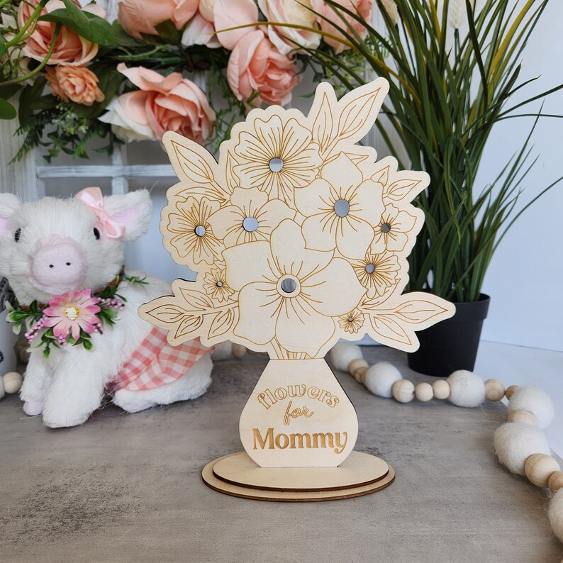 May include: A wooden floral bouquet decoration with the words "Flowers for Mommy" engraved on the vase. The bouquet features etched flower and leaf designs with small mirrored accents. The decoration sits on a round wooden base.
