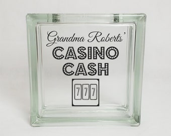 May include: A clear glass money jar with a black and white design that says "Grandma Roberts' Casino Cash" and has a slot machine graphic with the number 777.