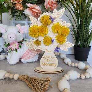 May include: A wooden floral arrangement with yellow dandelions and purple flowers. The vase-shaped base has the text "Flowers for Mommy". The arrangement is on a round wooden base. A stuffed pig and decorative garland are in the background.