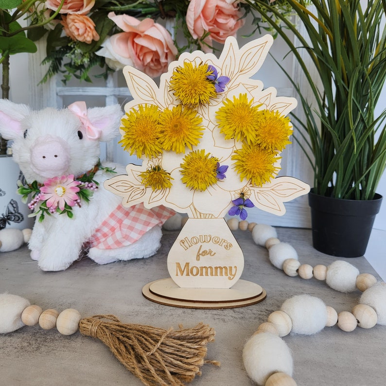 May include: A wooden floral arrangement with yellow dandelions and purple flowers. The vase-shaped base has the text "Flowers for Mommy". The arrangement is on a round wooden base. A stuffed pig and decorative garland are in the background.