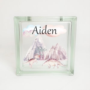 Puede incluir: Un bloque de vidrio con un dise&ntilde;o de acuarela de monta&ntilde;as, nubes y dinosaurios. El nombre "Aiden" est&aacute; escrito en letras negras en el bloque.