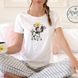 May include: A white t-shirt with a black and white cartoon cow wearing a sunflower on its head. The cow is standing on all four legs and has a pink nose.