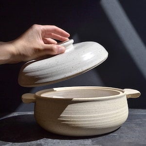 May include: A white ceramic pot with a lid. The pot has a speckled finish and a round shape. The lid is slightly domed and has a small knob on top.