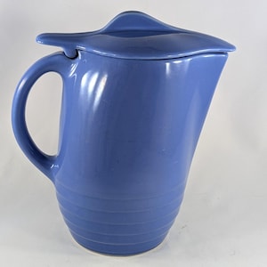 May include: A blue ceramic jug with a matching lid. The jug has a curved handle and a spout. The lid has a unique, stylized shape. The jug's body features horizontal ridges. This vintage piece is suitable for serving beverages.