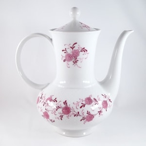 Può includere: Caffettiera in porcellana bianca con un motivo floreale rosa. La caffettiera ha un coperchio e un manico.