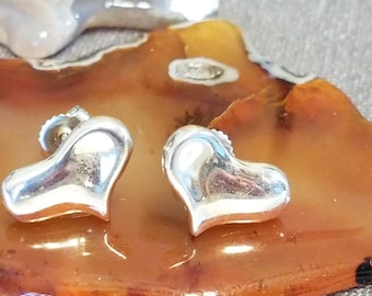 May include: A pair of silver heart-shaped stud earrings. The earrings are highly polished and reflect light. They are displayed on a brown and orange surface. The earrings are a romantic jewelry item.