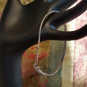 May include: A silver-colored chain bracelet with a textured, flat link design. The bracelet is displayed on a black mannequin hand against a colorful, patterned background. The clasp is visible, showing the bracelet's closure mechanism.