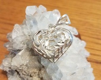 May include: A silver heart-shaped pendant with an open filigree design. The pendant is displayed on a cluster of clear quartz crystals, set against a wooden surface. The pendant's intricate details are visible.