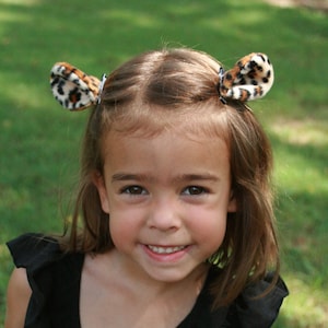 May include: A young girl with brown hair wearing a black sleeveless dress with ruffles. She is wearing leopard print hair clips on each side of her head.