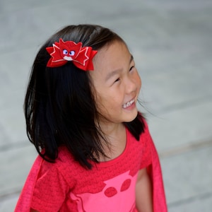 May include: A young girl wearing a red and pink superhero costume with a red bow in her hair. The costume has a pink owl logo on the front and a red cape.