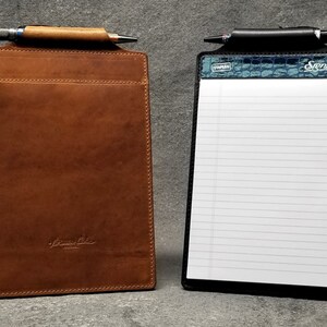 May include: Two leather clipboards, one brown and one black. The brown clipboard has a pen holder and a pocket. The black clipboard has a lined notepad and a blue and green crocodile pattern on the top. Both clipboards have a metal clip.
