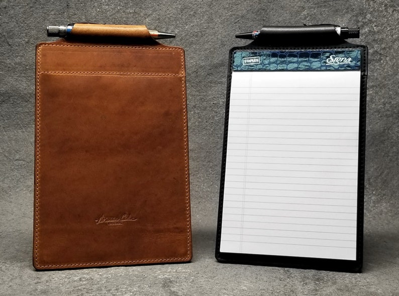 May include: Two leather clipboards, one brown and one black. The brown clipboard has a pen holder and a pocket. The black clipboard has a lined notepad and a blue and green crocodile pattern on the top. Both clipboards have a metal clip.
