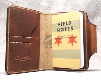 May include: A brown leather notebook cover with a pocket on the front. The cover is open to reveal a Field Notes memo book with the text "FIELD NOTES" and "48-Page Memo Book Durable Materials / Made in the U.S.A."