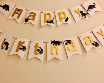 May include: A white banner with gold letters spelling "Happy Birthday". Black dinosaur silhouettes and gold letters are featured on the banner. The banner is strung with twine and is suitable for a birthday party.