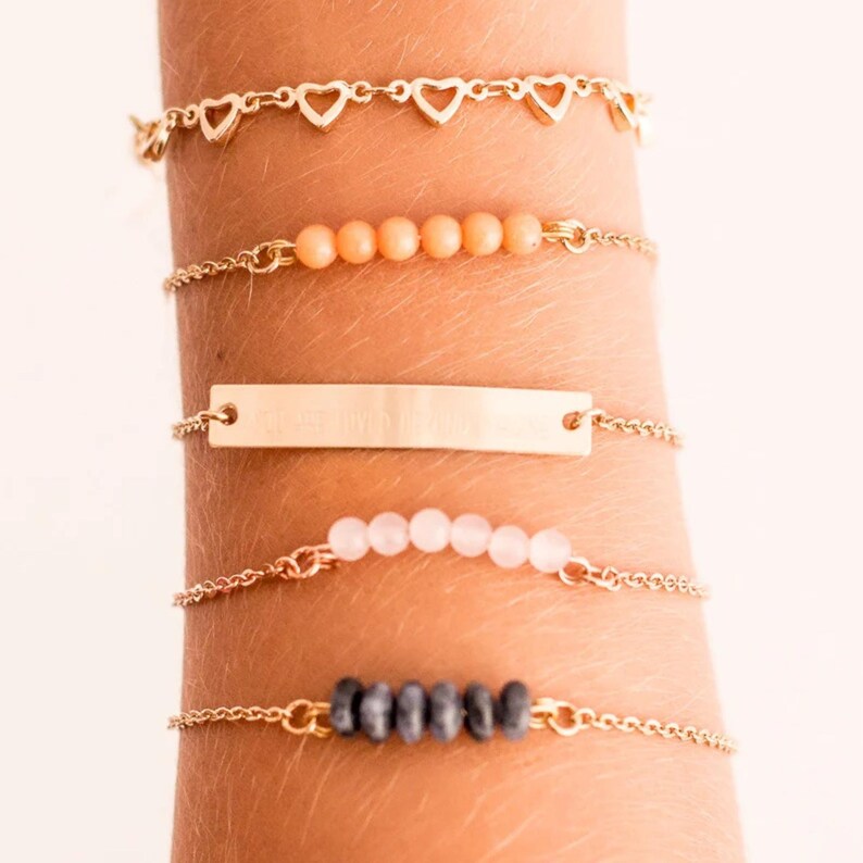 May include: Five gold-colored bracelets are displayed on a person's arm. The bracelets feature heart charms, peach-colored beads, a gold bar, pink beads, and dark gray beads. The bracelets are connected by delicate chains.