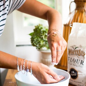 May include: A person's arms are adorned with delicate bracelets, some featuring charms and gemstones. The person is sprinkling flour into a white bowl. A bag of White Lily flour is visible in the background, suggesting baking or cooking.