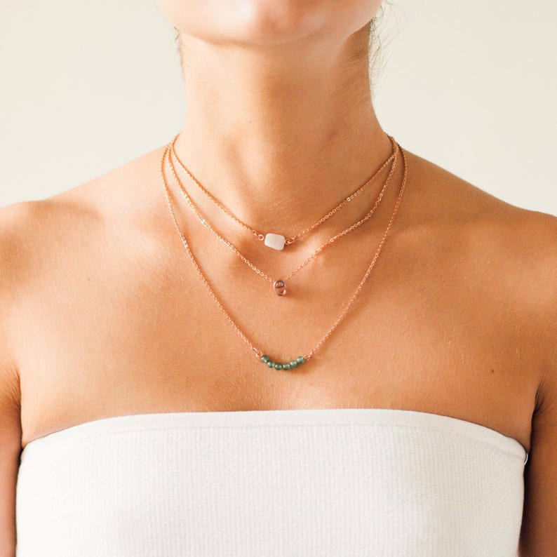 May include: Three layered necklaces with rose gold chains. The top necklace features a white rectangular stone, the middle one has a small pink pendant, and the bottom one has a row of green beads. The necklaces are displayed on a person.