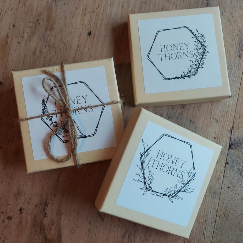 May include: Three small, beige square boxes with white labels featuring the text "HONEY THORNS" inside a geometric design with botanical illustrations. One box is tied with twine. The boxes are on a wooden surface.