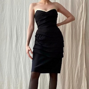May include: A strapless black dress with a sweetheart neckline and a cream-colored trim. The dress features a ruched bodice and a knee-length skirt. The model is wearing black tights and black heels.