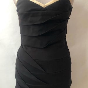 May include: A strapless black dress with a sweetheart neckline and a gold-colored trim. The bodice features horizontal pleats, and the skirt has diagonal pleats. The dress is displayed on a mannequin.