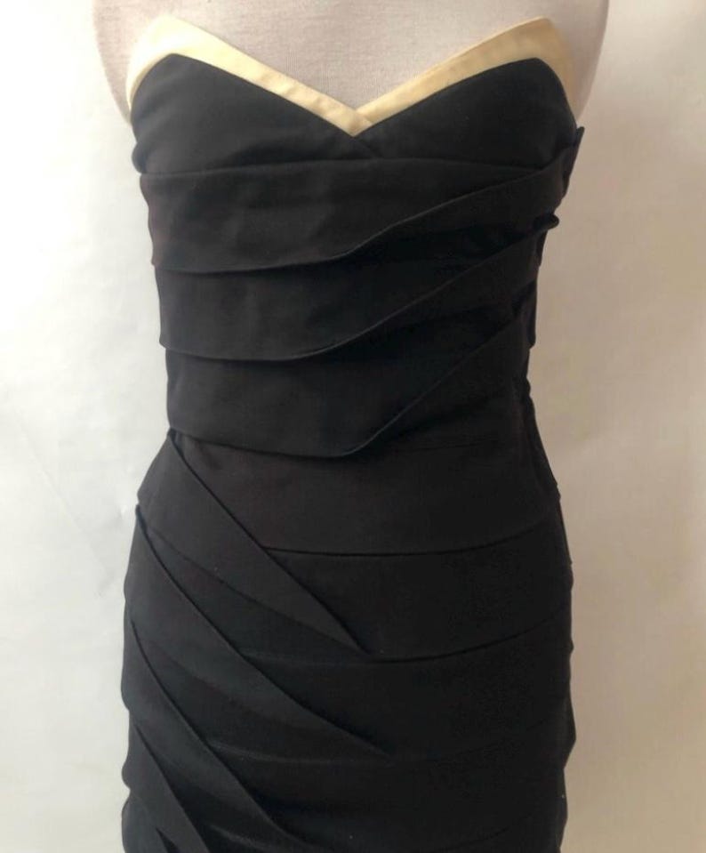 May include: A strapless black dress with a sweetheart neckline and a gold-colored trim. The bodice features horizontal pleats, and the skirt has diagonal pleats. The dress is displayed on a mannequin.