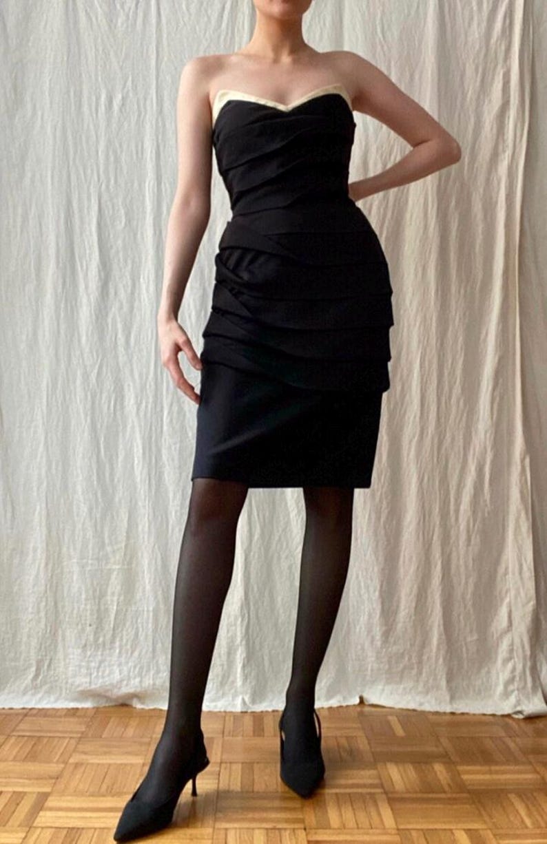 May include: A strapless black dress with a sweetheart neckline and a cream-colored trim. The dress features a ruched bodice and a knee-length skirt. The model is wearing black tights and black heels.