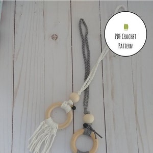 May include: Two wooden rings with crocheted tassels hanging from a cord. The ring on the left has a white tassel and the ring on the right has a grey tassel. The cord is made of grey and white yarn. The background is a white wooden surface. PDF CROCHET PATTERN is written on a white circle with green leaves.
