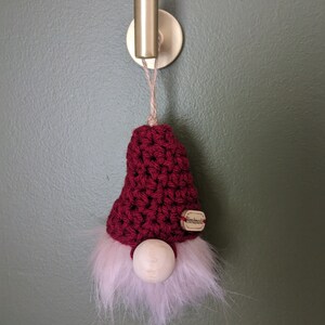 Könnte beinhalten: Ein roter Häkel-Zwergen-Anhänger mit einer Holznase und einem weißen, flauschigen Bart, der an einem goldenen Haken an einer grünen Wand hängt.