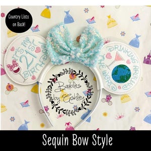 May include: A white headband featuring two circular ears with embroidery. The left ear reads It's my 21st Birthday with a wine glass icon. The right ear reads Drinking Around the World with a globe and martini glass. A light blue sequin bow sits between the ears.