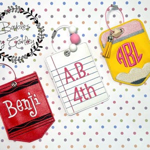 May include: Three personalized school-themed tags. One red crayon tag with the name "Benji". A white lined paper tag with "A.B. 4th" in pink. A yellow pencil tag with a pink monogram and a yellow tassel.