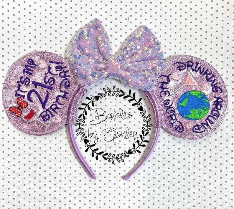 K&ouml;nnte beinhalten: Ein lavendelfarbenes Stirnband mit zwei runden Ohren aus violettem Glitzerstoff. Das linke Ohr zeigt den Text 'IT'S MY 21ST BIRTHDAY' mit einem Weinglas-Symbol. Das rechte Ohr zeigt 'DRINKING AROUND THE WORLD' mit einem Globus und einem Martiniglas.