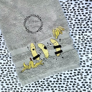 May include: A gray hand towel with embroidered bananas wearing sunglasses and a black band across their middle. The towel has the text "Bubbles by Bailey" in a circle.