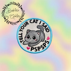 May include: Embroidered patch featuring a gray cat with large eyes and pink cheeks. The patch has the text "TELL YOUR CAT I SAID PSPSPS" in a circular design with a light blue border. Two pink hearts are also included.