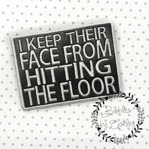 May include: A black rectangular patch with white embroidered text that reads "I KEEP THEIR FACE FROM HITTING THE FLOOR." The patch has a white border and is set against a white background with small gray polka dots. A black and white floral design is in the lower right corner.