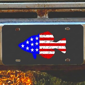 May include: Black license plate featuring a fish design in the colors of the American flag: blue, red, and white. The fish has a distressed, vintage look. The plate is mounted on a rusty vehicle bumper.