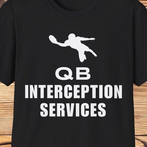 May include: Black t-shirt with a white silhouette of a football player throwing a pass. The text "QB INTERCEPTION SERVICES" is printed below the image.