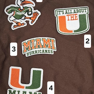 May include: Four embroidered patches on a brown background. Patch 1 shows a white ibis mascot in a green jersey. Patch 2 reads IT'S ALL ABOUT THE U. Patch 3 reads MIAMI HURRICANES. Patch 4 features a large U logo with the word MIAMI underneath.