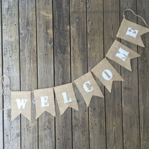 May include: A burlap banner with the word "WELCOME" spelled out in white letters. The banner is hanging on a wooden wall.