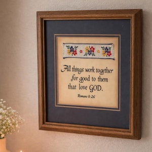 May include: Framed artwork with a wooden frame, featuring the quote "All things work together for good to them that love GOD." A floral embroidered banner is above the text. A lit candle and a vase of flowers are in the foreground.
