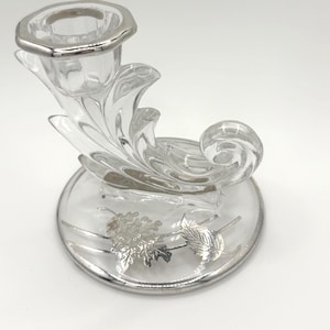 May include: Clear glass candlestick holder with silver-colored accents. The candlestick features a decorative, swirling design and a round base with silver leaf and floral details. The rim of the candle holder is also silver.