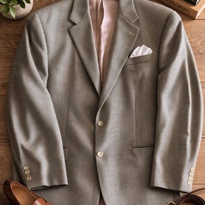 May include: A light gray blazer with a subtle checkered pattern, featuring a white pocket square. The jacket has two buttons and is displayed with brown leather shoes, a watch, and a belt. The blazer is hanging on a wooden hanger.