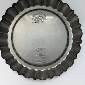 May include: A round, fluted metal tart pan with a silver finish. The pan has a scalloped edge and the text "Baker's Secret" and "Duncan Hines Tiara Desserts" are embossed on the surface.
