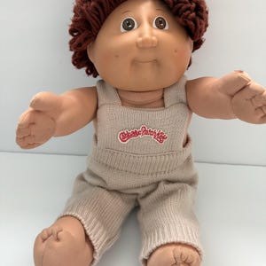 May include: A vintage Cabbage Patch Kid doll with brown hair and brown eyes. The doll is wearing a beige knit outfit with the Cabbage Patch Kids logo on the front. The doll is sitting with its arms outstretched.