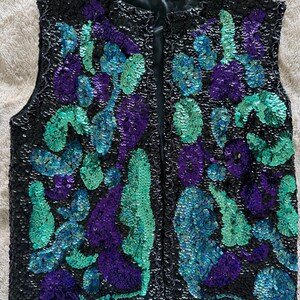 May include: A black sequined vest with a floral design in shades of purple, teal, and blue. The vest has a sleeveless design and a front closure. The sequins are densely packed, creating a shimmering effect. The vest is likely vintage or retro.